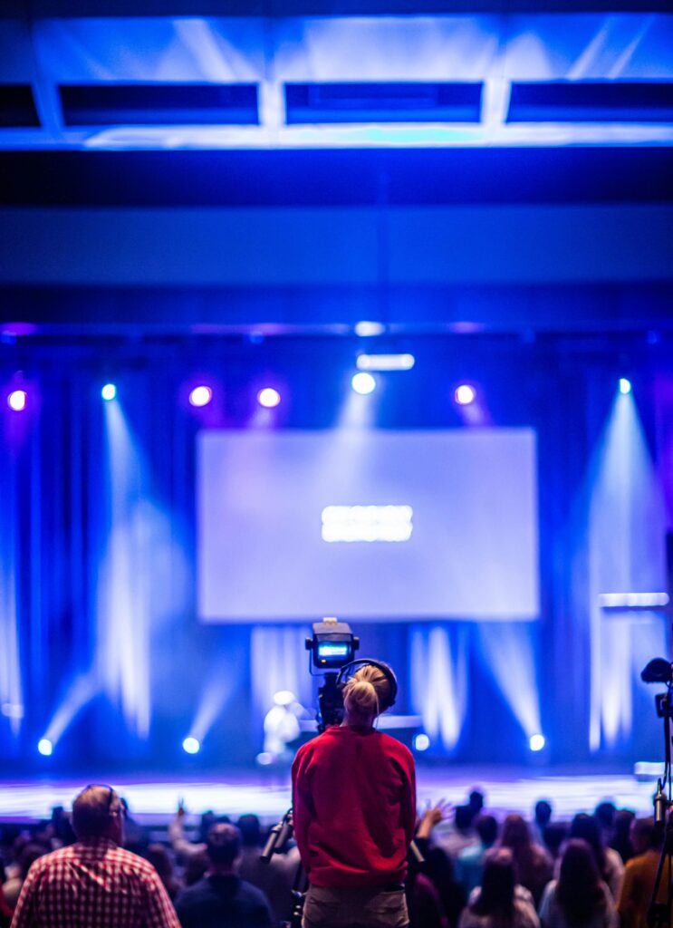 Cameraman capturing a live event on stage with colorful lights and blurred audience.